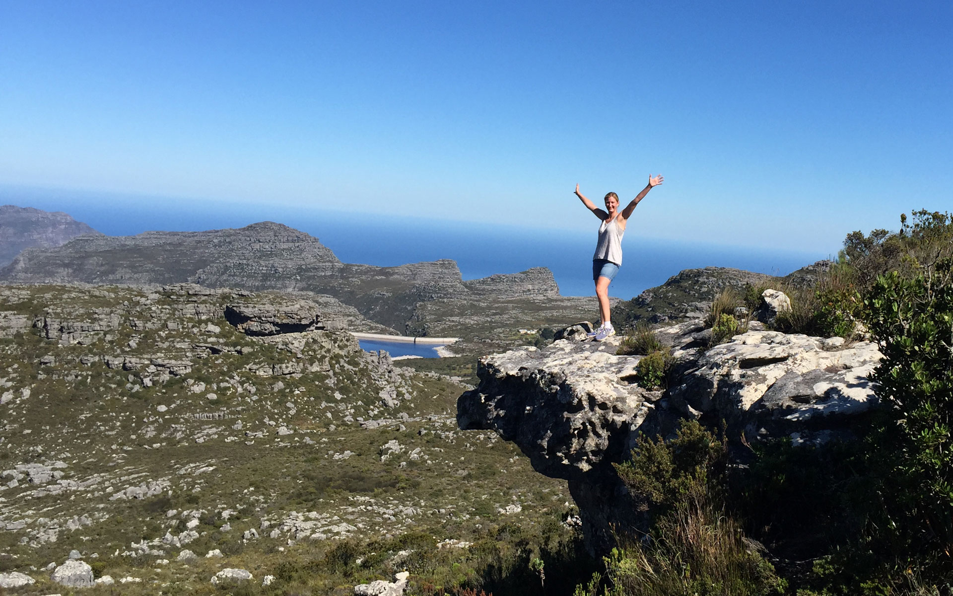 Reisebloggerin Barbara Freund steht auf einem Felsvorsprung des Tafelsbergs in Südafrika und reißt vor Freude die Arme in die Luft.