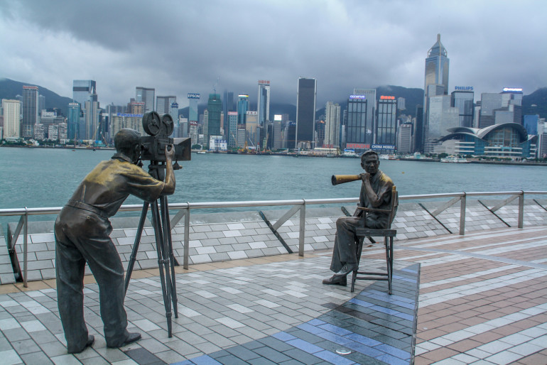 Zwei menschengroße Statuen bei Avenue of Stars stehen an der Ufernpromenade von Tsim Sha Tsuig mit Blick auf Hongkongs Skyline.