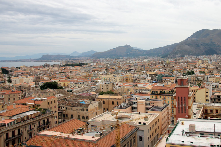 Bei einem Blick über die Dächer der Stadt Palermo sieht man im Hintergrund das Meer und die Berge.