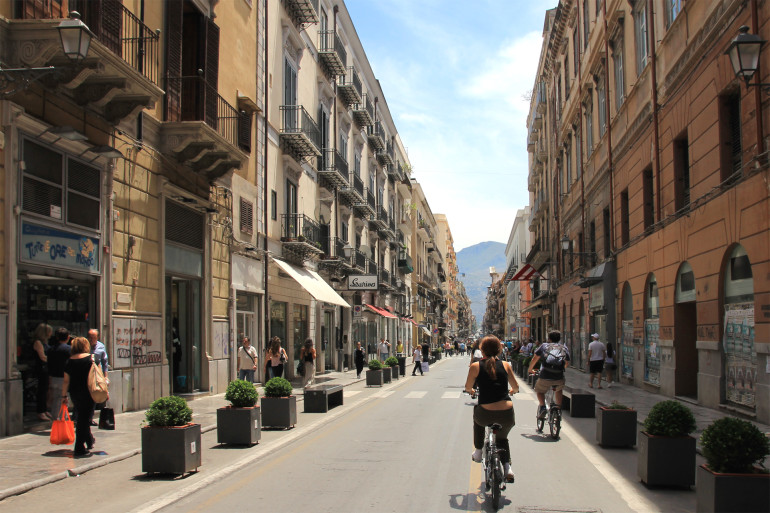 Zwischen barocken Häuserfassaden zeigt sich Palermos Fußgängerzone Via Maqueda mit ihren Besuchern, am Ende der Straße erblickt man einen Berg.