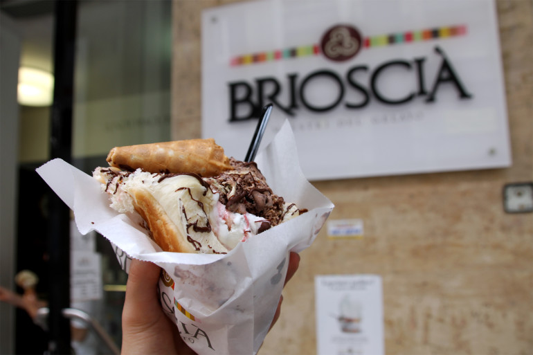 Vor dem Schild der Eisdiele Briosciá in Palermo hält die Bloggerin ein Portion Eis im Brötchen in die Kamera.