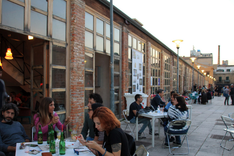 Vor der langen Fensterfront des Restaurants Cantieri Culturali alla Zisaeines auf einem ehemaligen Industriegelände in Palermo sitzen Gäste an Tischen, essen, trinken und unterhalten sich.