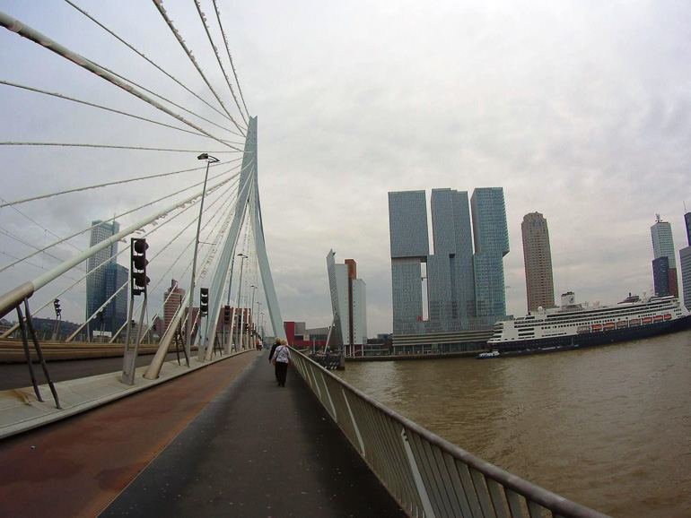 Eine Eisenstahl-Brücke in Rotterdam führt über das Wasser, auf das ein Schiff fährt, zu modernen Wolkenkratzern.