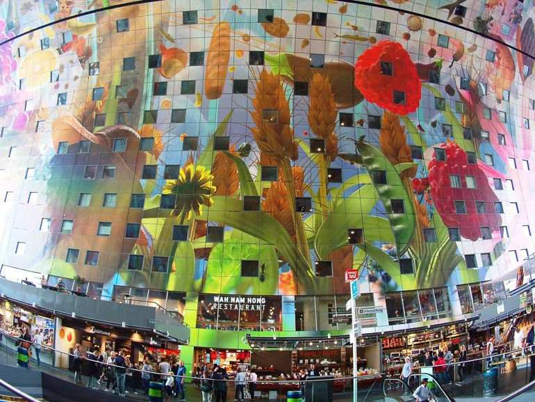 Das Rotterdamer Markthal von innen beherbergt eine riesige Glasfront mit aufgezeichneten Blumen und vielen verschiedenen Verkaufsläden, an denen sich Menschen tummeln.
