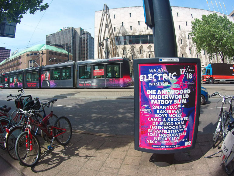 Auf einer Straße in Rotterdam wurde ein Musikfestival-Plakat angebracht im Hintergrund fährt die Straßenbahn entlang der Hochhäuser.