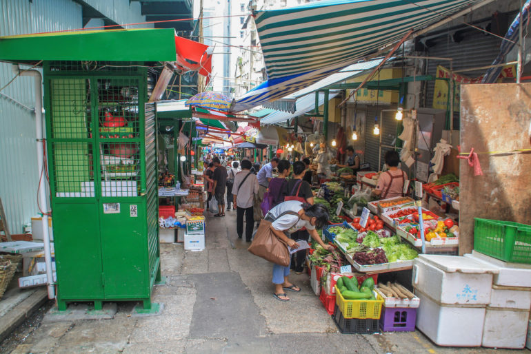 In einer schmalen Gasse in Hongkong tummeln sich Einheimische auf einem Markt, die an Ständen unter Markisen Obst und Gemüse verkaufen.