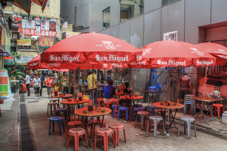 In einer Straße in Hongkong steht eine Garküche mit roten Sonnenschirmen und Plastikstühlen.