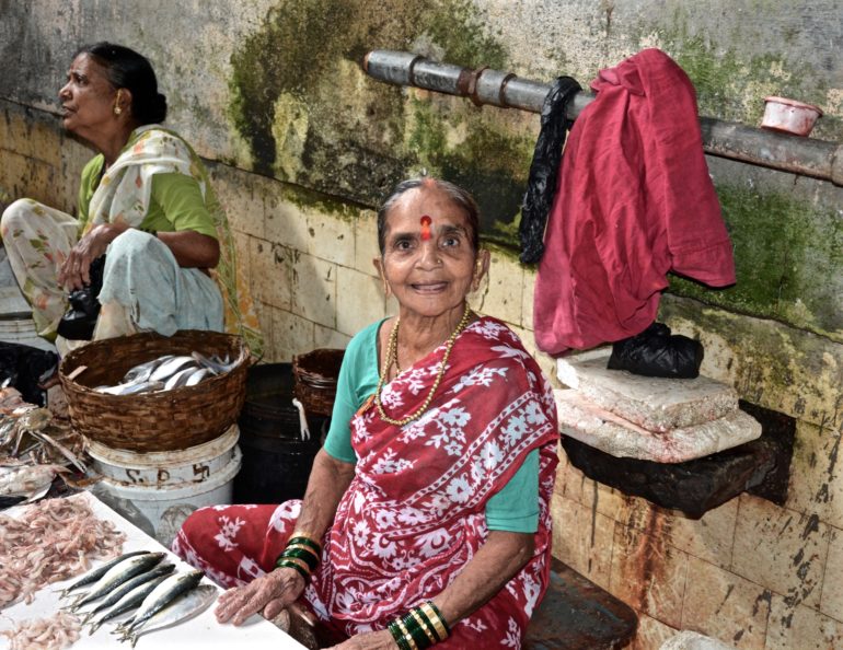 In Mumbais Central Fischmarkt Koli sitzt eine einheimische Frau im Schneidersitz vor ihrem Verkaufstisch mit Fisch und lächelt in die Kamera.