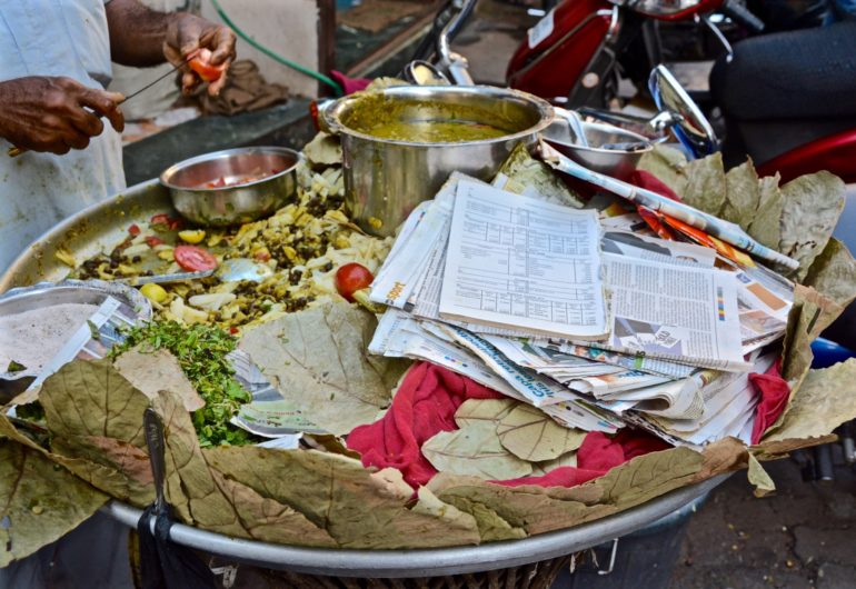 Ein Einheimischer schneidet über einer großen silbernen Schüssel eine Tomate, die Schüssel ist mit Töpfen indischer Streetfood-Spezialitäten, Blättern und Zeitungen zum Einwickeln gefüllt.