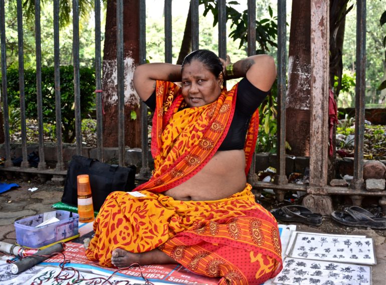 Auf einer Straße im Mahalaxmi Viertel von Mumbai sitzt eine einheimische Frau vor einem Eisenzaun und bietet vorbeilaufenden Passanten Henna Tätowierungen an.