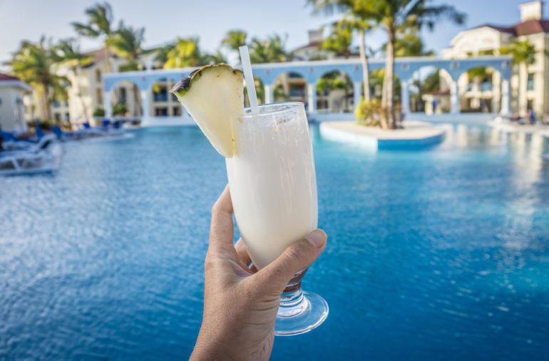 Eine Ananas verziert das in die Kamera gehaltene Cocktailglas gefüllt mit Piña Colada, im HIntergrund erstreckt sich die Poollandschaft eines Hotels.