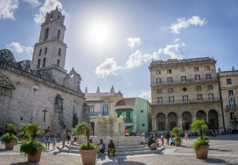 Blick auf die von der Sonne angestrahlte Häuserfassaden, auf der linken Seite ist eine typische kubanische Kirche zu sehen und in der Mitte des Platzes tummeln sich Menschen an einem Brunnen.