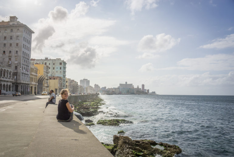 Eine Frau sitzt in Malecón am Straßenrand, welcher an der Küste zum Meer liegt und lächelt in die Kamera.