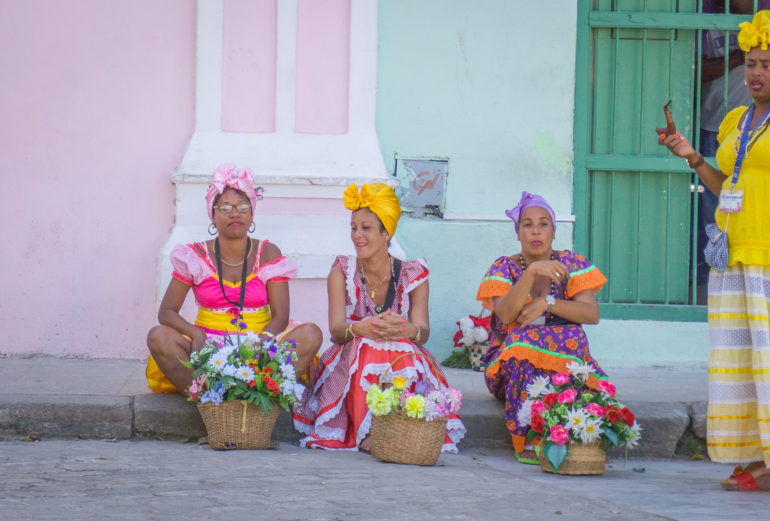 Drei Frauen in bunten kubanischen Gewändern sitzen mit ihren Blumenkörben an einem Straßenrand in Havanna, eine Frau in gelbgekleideter Tracht gesellt sich zu ihnen und raucht Pfeiffe.