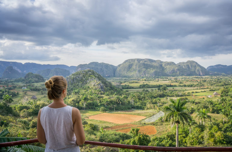 Eine Frau blickt mit dem Rücken zur Kamera auf die mit Palmen und Hügeln umgebene Landschaft Viñales in Kuba.