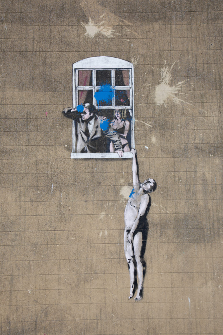 An einer Häuserfassade in Bristol ist ein Bild in Banksys Stil zu sehen - ein Paar gezeichnet, bei dem ein Mann mit Fernglas aus dem Fenster blickt, während die Frau besorgt in Unterwäsche an seinem Arm greift, während ein weiterer Mann sich mit einem Arm nackt am Fenstersims festhält.