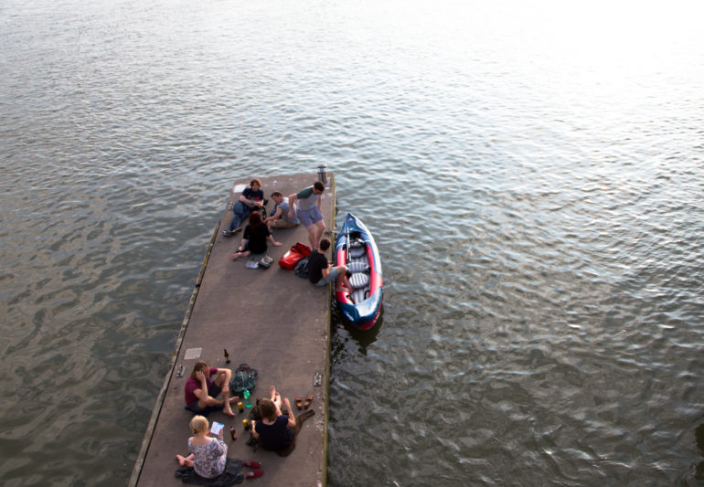 Auf einem Steg am Kanal von Bristol sitzen junge Menschen und unterhalten sich, an der Seite des Stegs sind gerade zwei Personen dabei in ein Kajak zu steigen.