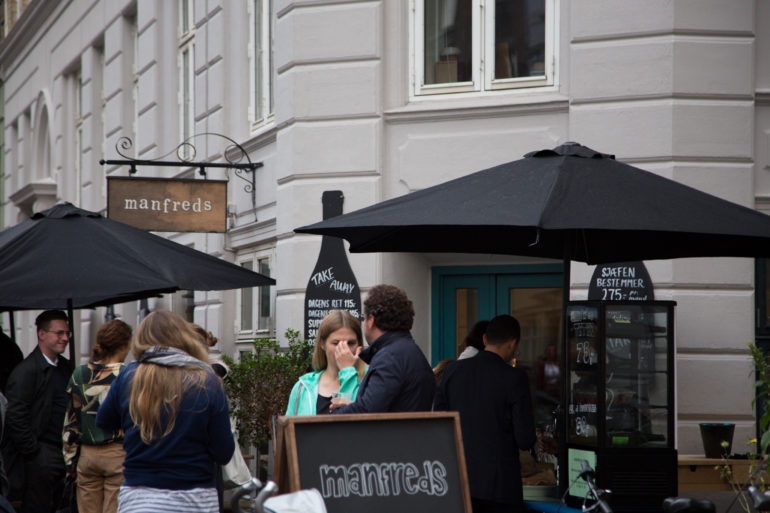 Vor dem Eingang der Bar manfreds in Kopenhagen haben sich Menschen unter scharzen Sonnenschirmen gestellt und unterhalten sich.