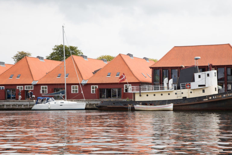 Ein Segelschiff und ein Schiff haben vor einem Ufer in Kopenhagen geankert, im Hintergrund stehen rote Holzhäuschen mit weißen Fenstern.