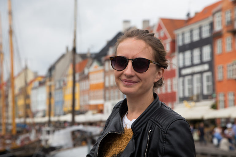 Die Bloggerin Anika lächelt mit ihren braunen Haaren und einer schwarzen Sonnenbrille und Lederjacke in die Kamera, im Hintergrund ist unscharf die Häuserfassade der Innenstadt von Kopenhagen zu erkennen.