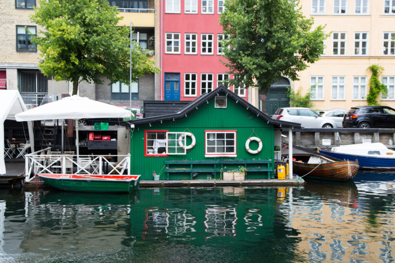 An einem Kanal in Kopenhagen steht ein grünes Hausboot mit kleiner Terrasse auf der ein weißer Sonnenschirm gespannt ist, an der Hausfassade hängen zwei Rettungsringe.