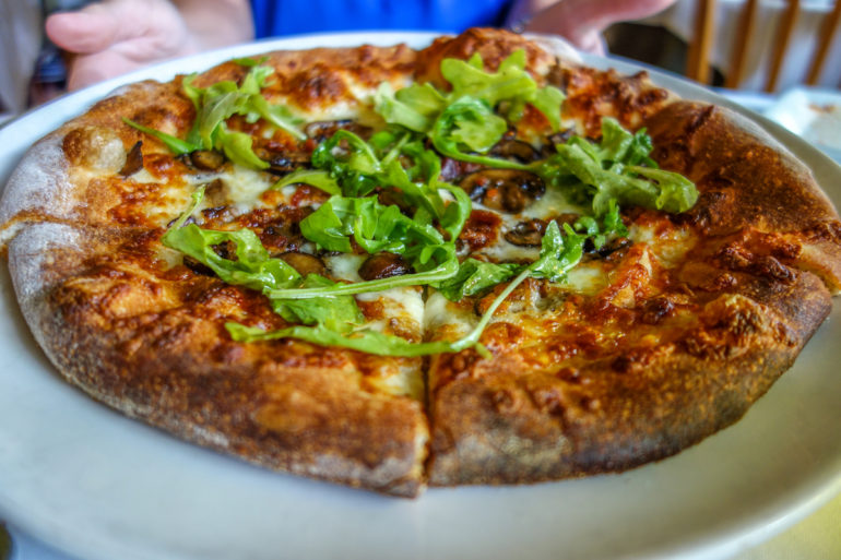 Zwei Hände halten in Virginia im Restaurant Magnolias Pizza einen Teller mit einer knusprigen Pizza belegt mit Pilzen und Ruccola in die Kamera.