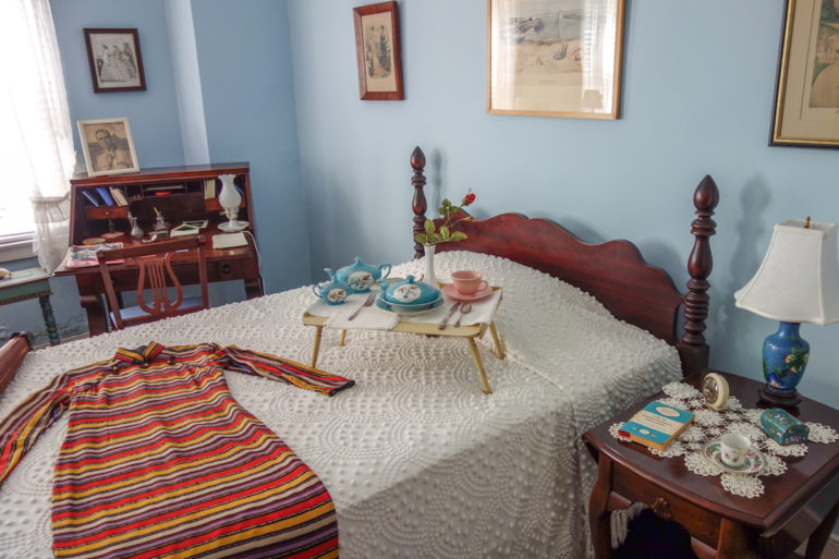 In einem hellblauen Schlafzimmer in Virginias Marshall House steht ein Sekretär, ein Bett und ein Nachttisch in dunklem Holz, auf dem Bett wurde auf der weißen Überwurfdecke ein buntgestreiftes Kleid und ein Frühstücksbrett mit hellblauem und rosafarbenen Geschirr drapiert.