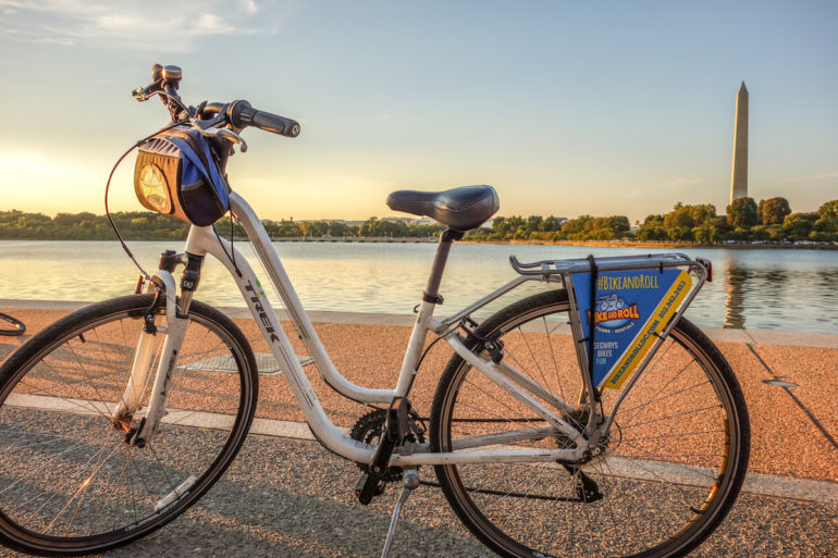 Ein weißes Leihfahrrad von Bike and Roll steht in Washington D. C., USA auf einer Straße, im Hintergrund ist das Washington Monument und Wasser bei Sonnenuntergang zu sehen.