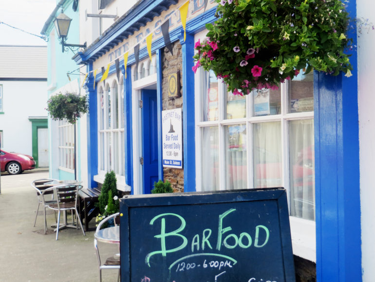 Im kleinen irischen Örtchen Schull ist der Blick auf eine Straßenzeile mit einem weiß und blau getünchtes Pub mit Blumen und Wimpeln gerichtet.