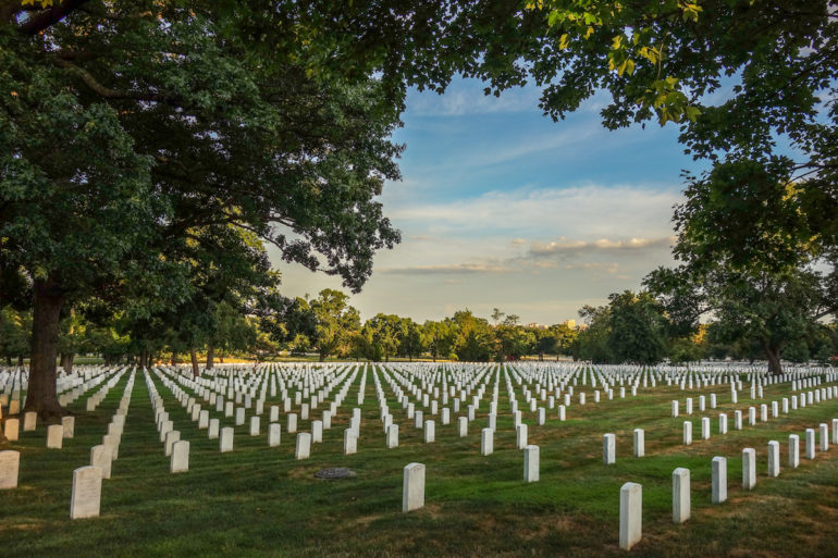 Auf dem Friedhof Arlington Cemetery in Virginia stehen auf einer Wiese umzäunt von hohen Bäumen in Reihen weiße Grabsteine.