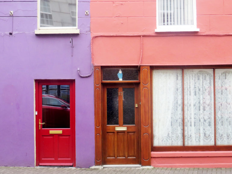 Der Blick auf eine Häuserfassade in Clonakilty, Irland, zeigt eine lila Wand mit roter Tür, sowie nebenan eine rosa Hauswand mit dunkelbrauner Holztür und Fenstern