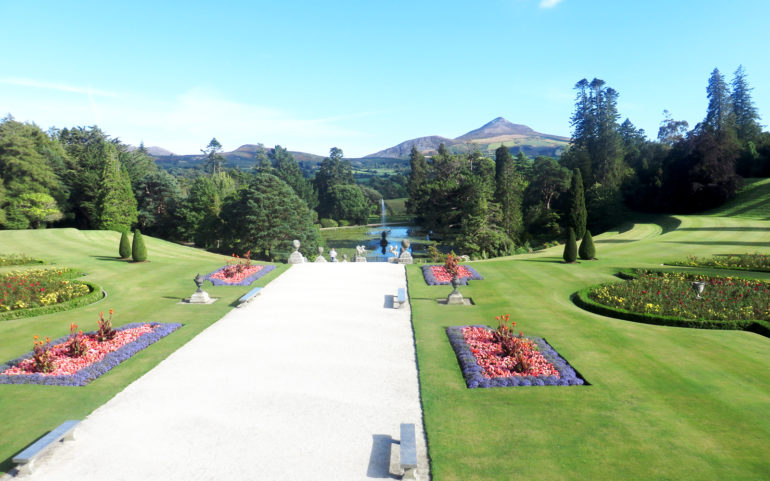 Ein heller Kiesweg mit Sitzbänken führt durch die Powerscourt Gardens zum See, im Hintergrund sind die umliegenden Hügel zu sehen.