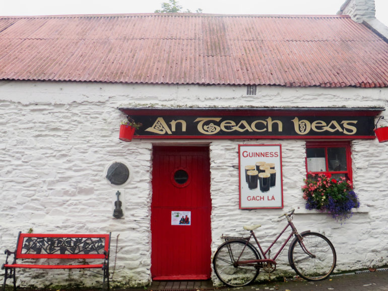 An das idyllisch wirkende Haus des Pubs "An Teach Beag" in Clonakilty, Irland, ist ein Fahrrad angelehnt, eine rote Bank ist farblich an die Tür und das mit Blumen beschmückte Fenster angepasst.