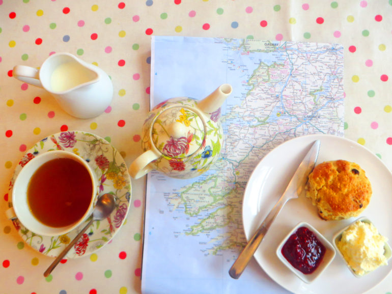 Auf einer bunt gepunkteten Tischdecke im Gallery Café in Irland steht auf einer ausgeklappten Landkarte eine Tasse Tee mit Kännchen, sowie auf einem Teller angerichtet ein Scones mit Butter und Marmelade.