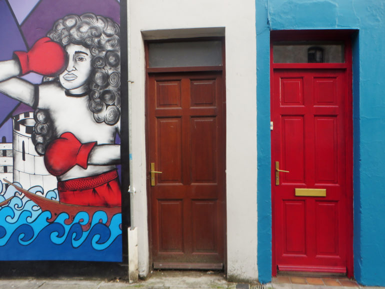 Neben zwei bunten Türen in Cork wurde an die Hausfassade eine lockige Boxerin mit roten Handschuhen gezeichnet.