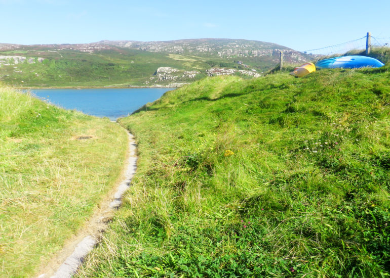 Der Weg zur Bucht von Crookhaven in Irland führt an grünen Wiesen vorbei zum Meer, im Hintergrund sind bewachsene Hügel zu sehen.