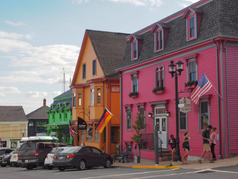 Die Fassaden der Holzhäuschen entlang einer Straße in Lunenburg sind in farbenfrohes rosa, orange und grün gestrichen, Autos parken davor und an den Häusern hängen unterschiedliche Länderfahnen.