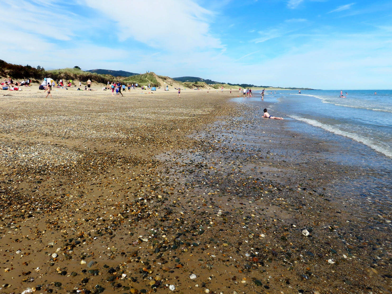 Das Meer am Strand von Brittas Bay, südlich von Wicklow, hat viele kleine Muscheln und Steine angespült, im Hintergrund tummeln sich Badegäste.