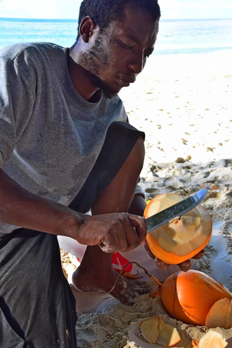 Auf den Seychellen: Ein einheimischer Mann schlägt eine orangene Kokosnuss mit einem Messer am Strand von Beau Vallon auf.