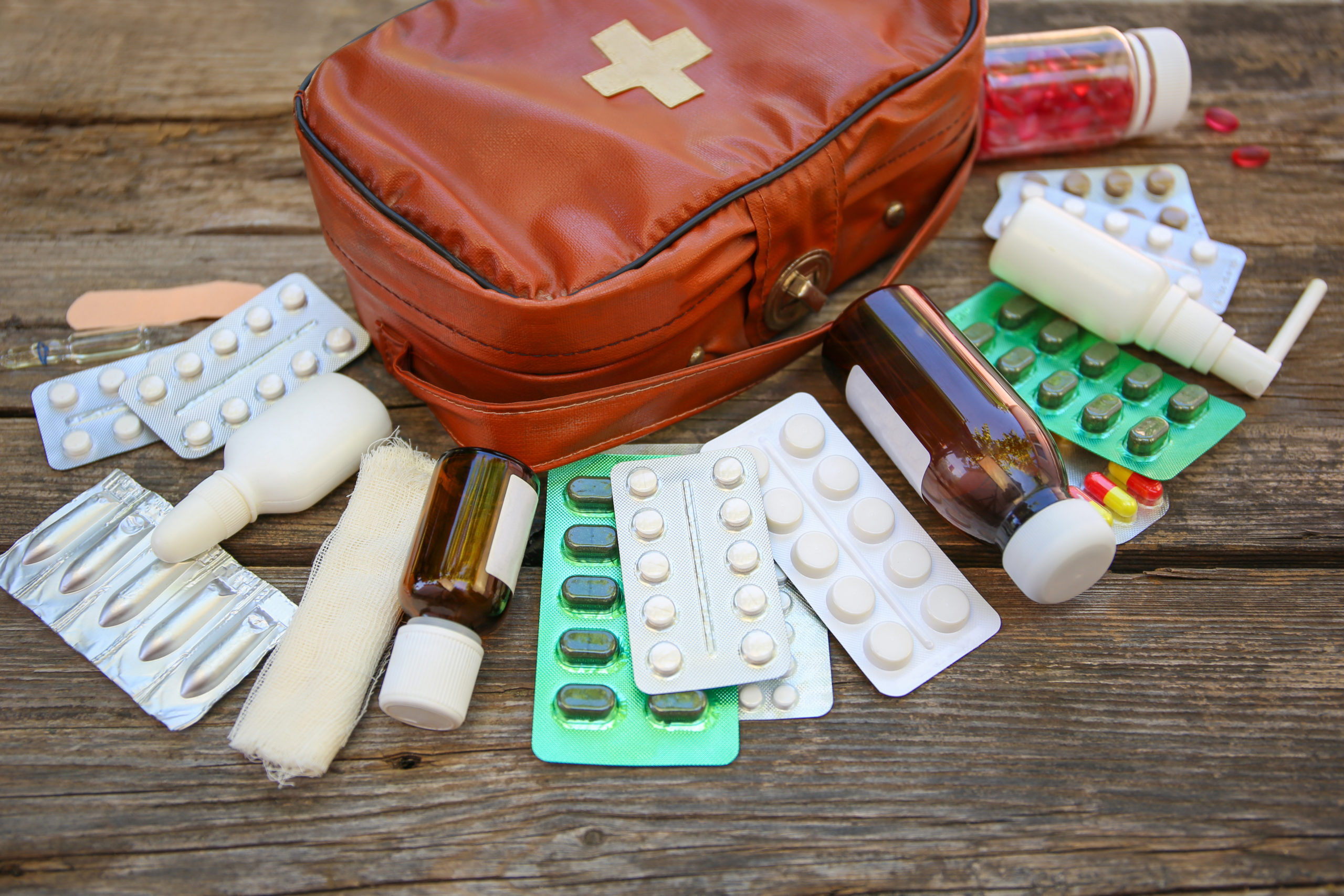 Eine Reiseapotheke in Form einer Erste-Hilfe-Tasche liegt auf Holz, davor liegen Tabletten, Zäpfchen, Medikamentenflaschen und Verbandszeug.