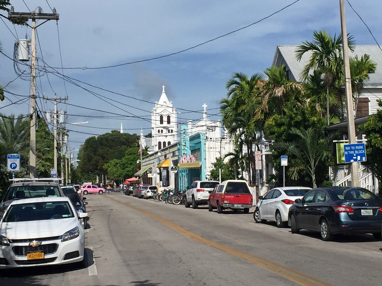 Einige Autos parken entlang einer Straße von Key West, USA, auf der rechten Straßenseite sind kleinere Läden und eine weiße Kirche.