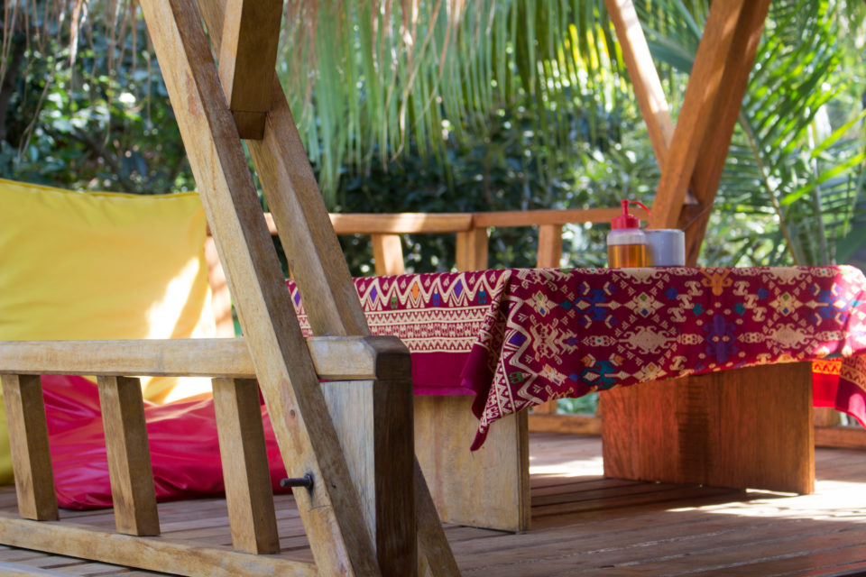 Eine hölzerne Sitzecke mit bunten Stoffen auf einer Terrasse eines Cafés inmitten tropischer Pflanzen lädt zum Verweilen ein.