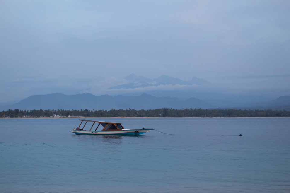 Auf dem Meer der Insel Gili Air auf Indonesien schippert einsam ein Fischerboot im Hintergrund ist durch feinen Dunst die Insel Lombok im Umriss zu erkennen.