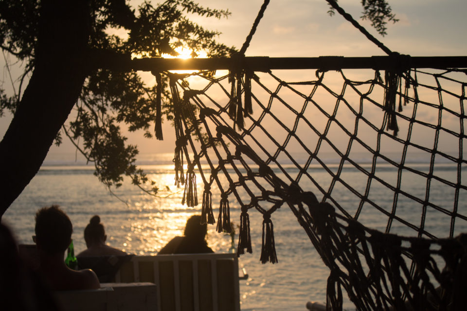 Vor einer gespannten Hängematte des Casa Vintage Beach Restaurants auf Gili Trawangan im Westen der Gili Inseln sitzen Menschen am Strand und beobachten den Sonnenuntergang.