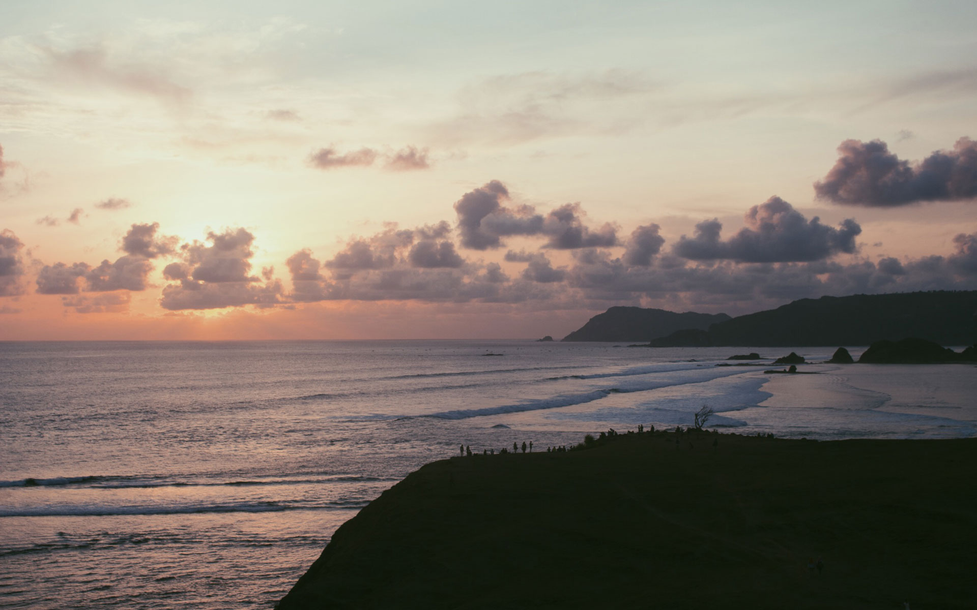 Am Strand von Tanjung Aan in Lombok, Indonesien, geht über dem Meer die Sonne unter, dessen Wellen sanft auf die bewachsenen Hügeln schlagen.