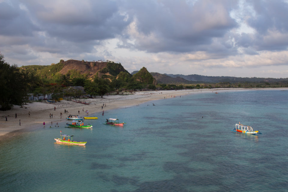 Fischerboote und Badegäste tummeln sich am Ufer des Tanjung Aan Beaches vor den dichtbewachsenen Hügeln der Insel Lombok.
