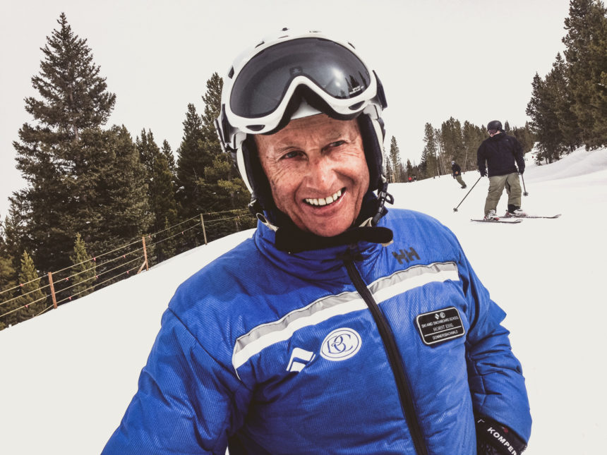 Ein 77jähriger Skilehrer mit blauer Jacke, Helm und Skibrille lächelt an der Skipiste von Vail, USA in die Kamera.