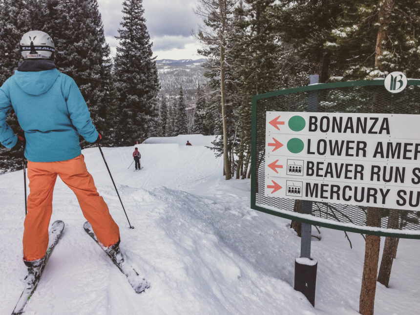 Eine Skifahrerin mit Helm, blauer Jacke und orangefarbener Hose steht neben einem Hinweisschild zu Beginn der Buckelpiste Bonanza bei Breckenridge, USA und blickt pistenabwärts.