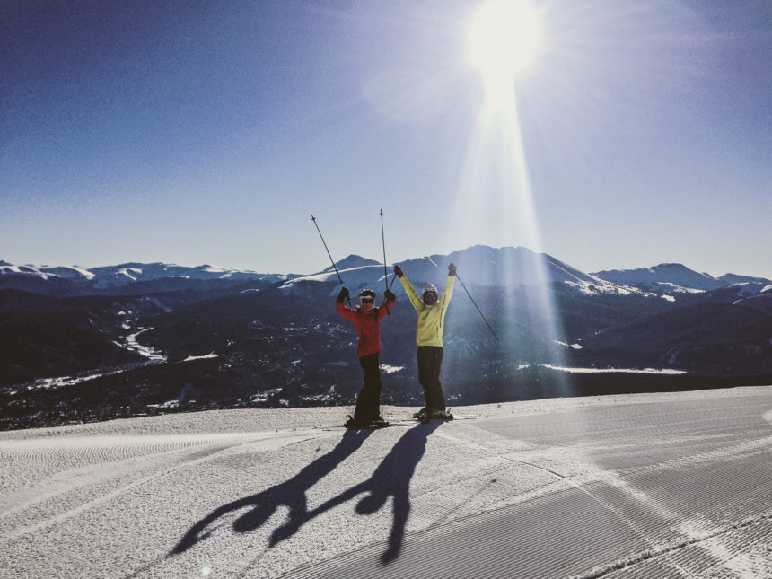 Zwei Skifahrer stehen vor einer Bergkulisse bei strahlendem Sonnenschein auf der Skipiste von Breckenridge und halten ihre Skistöcke in die die Luft.