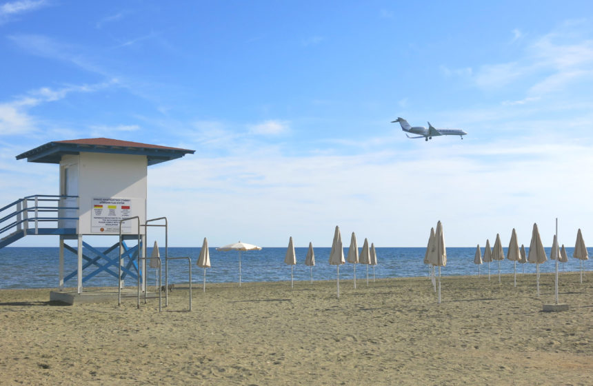 Über den menschenleeren Sandstrand Makenzy und seinem Lifeguard-Häuschen und weißen Schirmen ist ein Flugzeug im Landeanflug auf Zyperns Flughafen Larnaka..